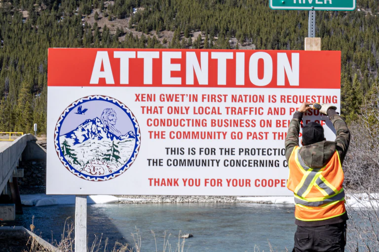 Man stands next to sign that reads 'Attention: Xeni Gwet'in First Nation is requesting that only local traffic and persons conducting business on behalf of the community go past this point'