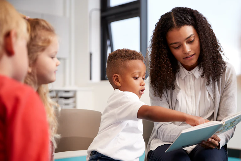 A child aged about two and a half points to a book held by a teacher in a circle of children.