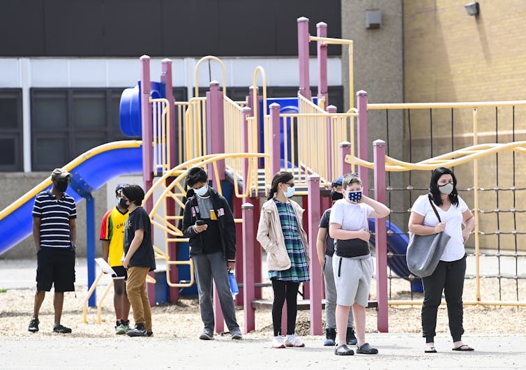 Parents and young youth wearing masks stand in a line in a playground.