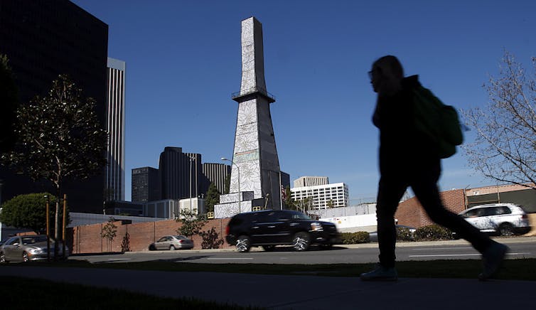 A silhouetted student with a backpack walks past an oil derrick covered with drawings of flowers outside a school.