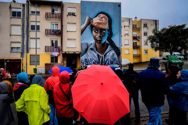 A crowd of people in rainy weather stand and look at a mural that covers a wall of a building. It depicts a dark-skinned woman lifting a white-skinned mask off her face.