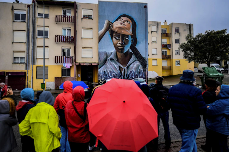 A crowd of people in rainy weather stand and look at a mural that covers a wall of a building. It depicts a dark-skinned woman lifting a white-skinned mask off her face.