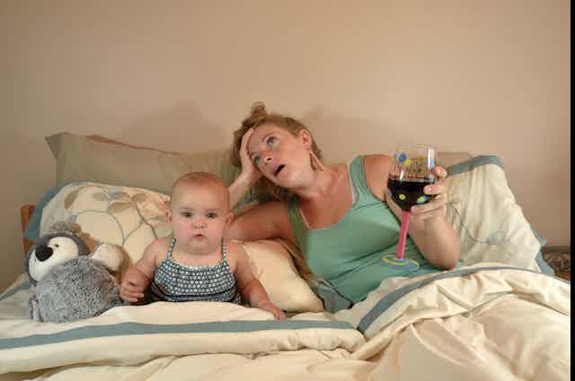 Mom lays in bed with baby. Mom has glass of wine in hand, looks exhausted
