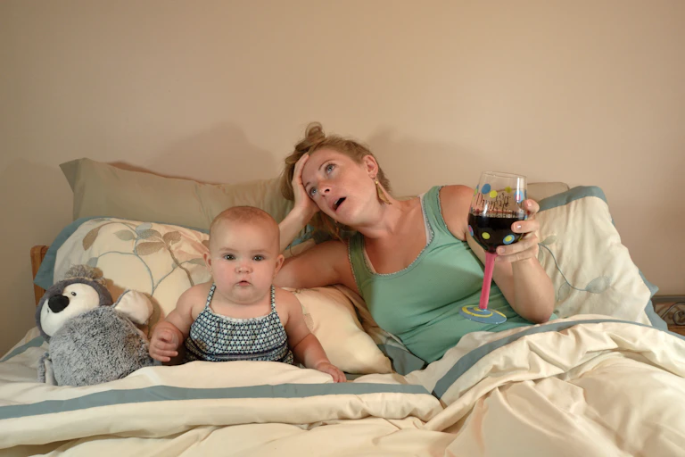 Mom lays in bed with baby. Mom has glass of wine in hand, looks exhausted