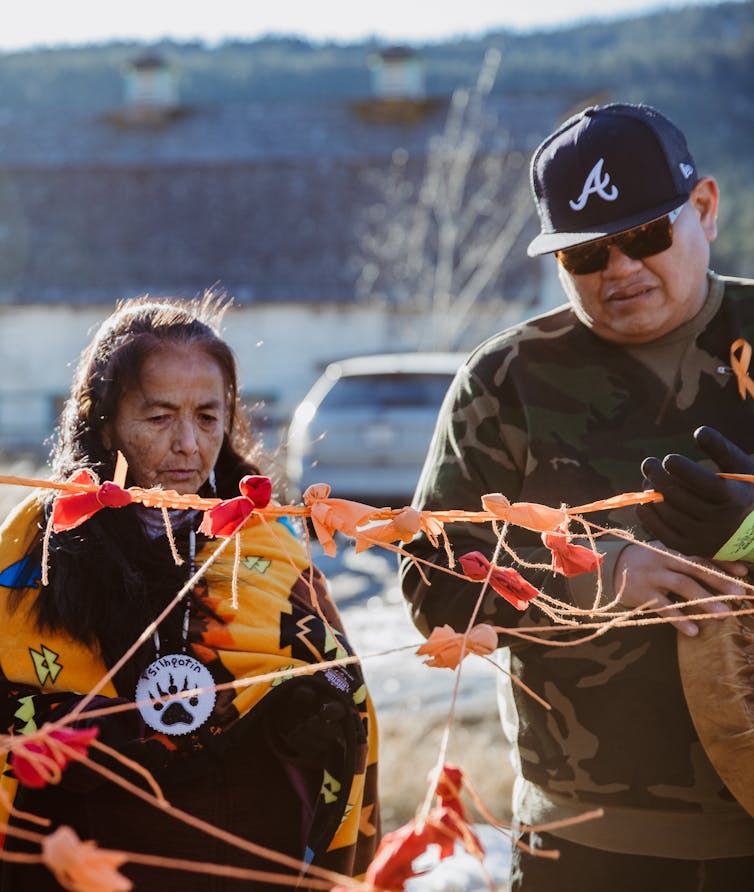 Two Indigenous people (one male, one female) stand around orange ribbon and orange ties.