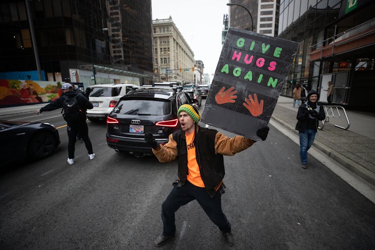 Man holds sign in traffic that reads 'Give hugs again'