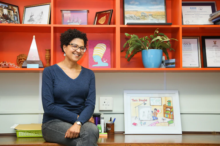 A smiling woman wearing a blue shirt and dark jeans is perched on the edge of a desk. Behind her is an orange shelf displaying various objects and items.