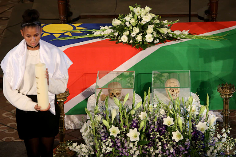 A girl holds a large candle near two human skulls encased in glass.