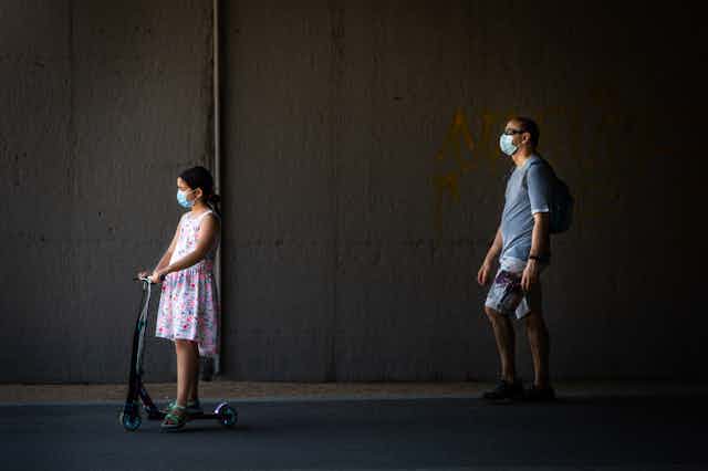 Masked father walks behind masked daughter riding scooter