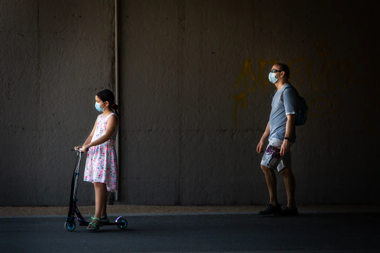 Masked father walks behind masked daughter riding scooter