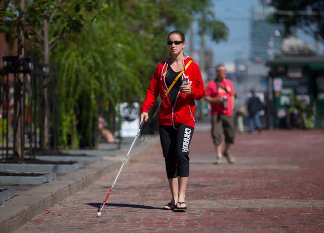 A woman walks on a brick road in a city wearing sunglasses and holding a white cane in her right hand and a smart phone in her left hand