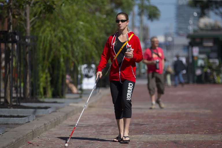 A woman walks on a brick road in a city wearing sunglasses and holding a white cane in her right hand and a smart phone in her left hand