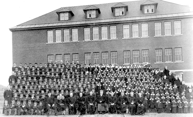 A black and white photo of dozens of Indigenous boys and girls lined up in front of the school while a row of church and school officials sit in the front of the picture.