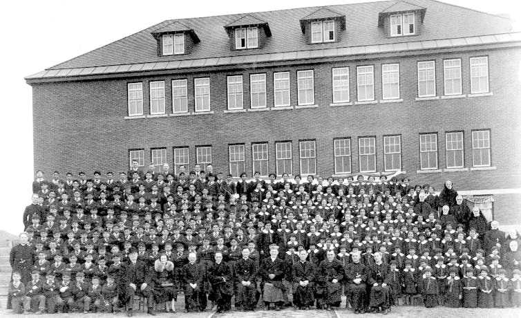 Uma foto em preto e branco de dezenas de meninos e meninas indígenas alinhados em frente à escola, enquanto uma fileira de funcionários da igreja e da escola estão sentados na frente da foto.