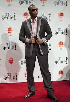 Kardinal Offishall, in a suit and pink shirt, poses on the red carpet.