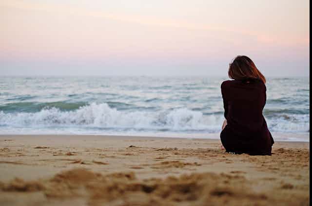 A woman sits on the beach, facing the ocean.