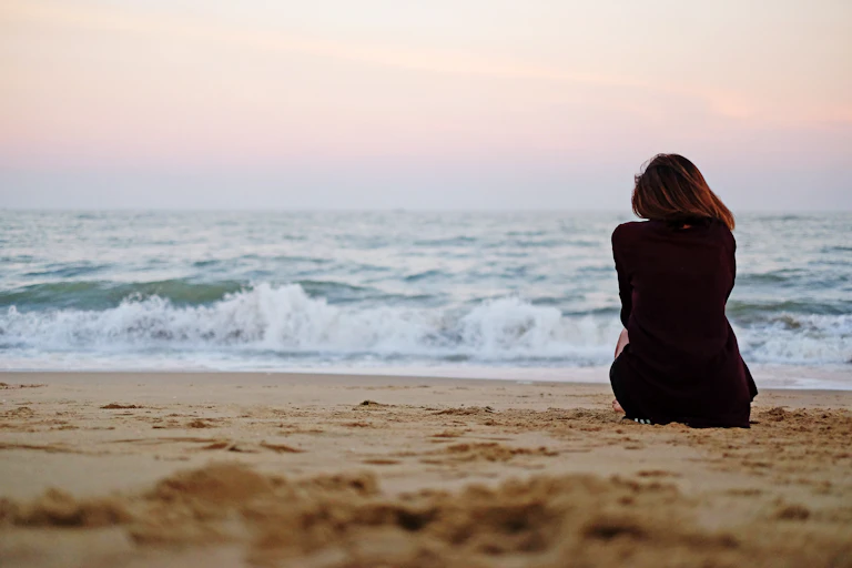 A woman sits on the beach, facing the ocean.