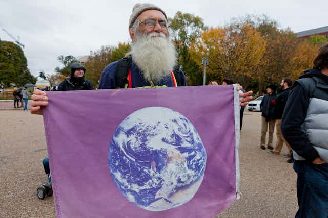 Old man with large white beard holds flag with image of Earth from space