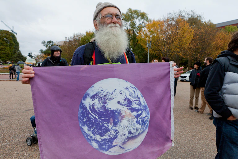 Old man with large white beard holds flag with image of Earth from space