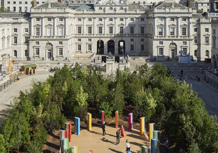 Circle of coloured monoliths surrounded by trees in front of large neoclassical building.