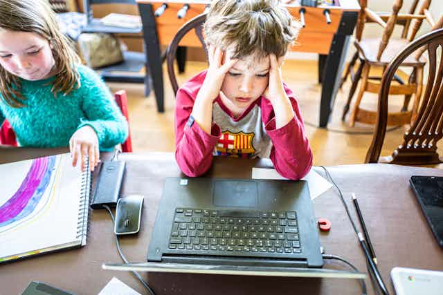 A little boy in a red t-shirt holds his head in his hands as he does an online school lesson from home with his sister
