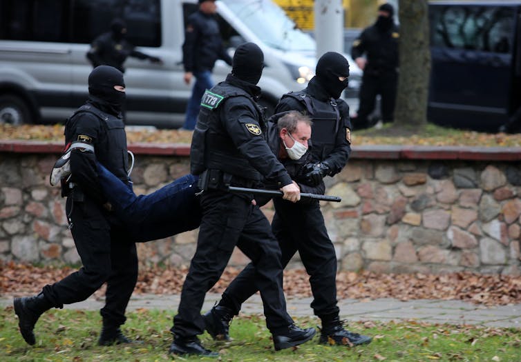 Police in riot gear and holding batons physically carry a man down the street