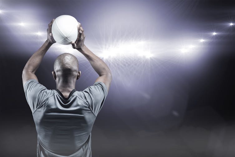 Back of man facing stadium lights and holding rugby ball over his head