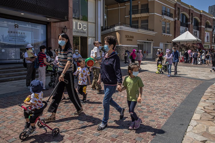A woman pushes a child on a stroller, while walking beside an older woman holding the hand of a young girl. They are walking past shops at an outdoor shopping mall.