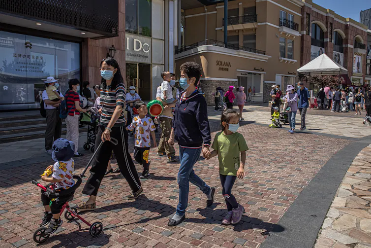 A woman pushes a child on a stroller, while walking beside an older woman holding the hand of a young girl. They are walking past shops at an outdoor shopping mall.