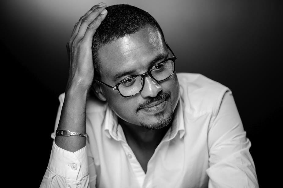 A black and white image of a man in spectacles, short hair and a white button-down shirt. He has a hand on his head and a wry smile.