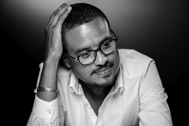 A black and white image of a man in spectacles, short hair and a white button-down shirt. He has a hand on his head and a wry smile.
