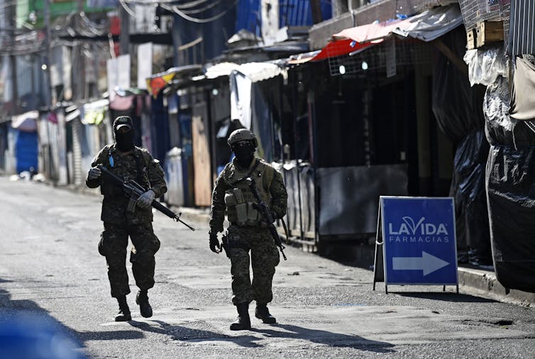 Two armed soldiers in full fatigues and face masks walk down a street lined with vendors