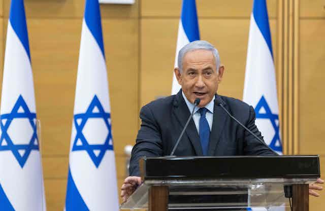 Benjamin Netanyahu speaks at a podium in front of four Israeli flags.