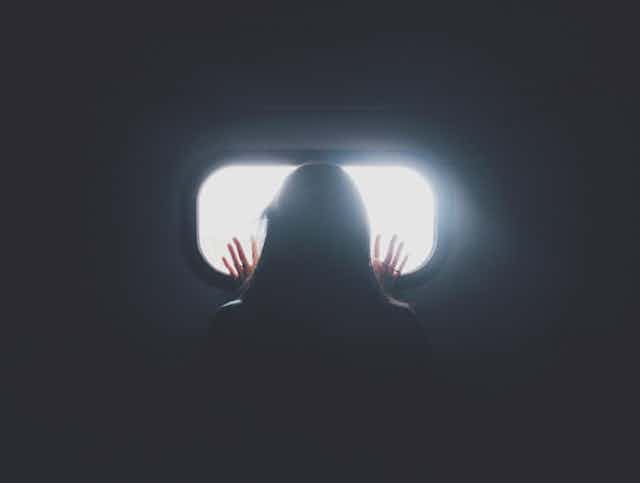 Woman stares looking out a small window in a dark room