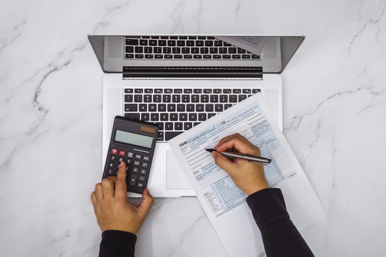 Someone's left hand is holding a calculator while the right hand is holding a pen on a 1040 form, atop a laptop