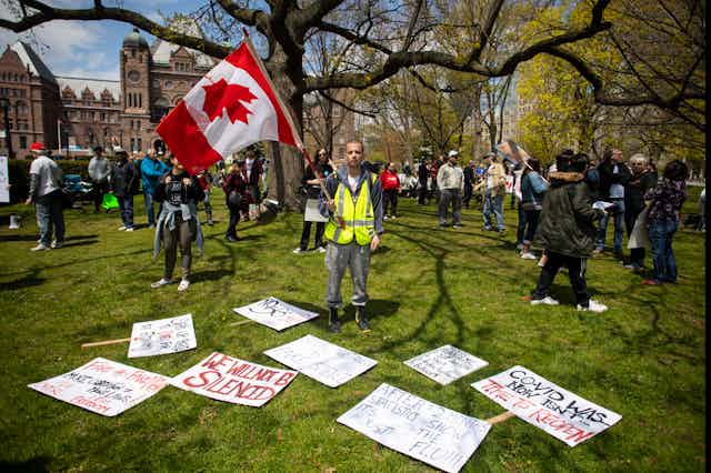 (Man stands holding an upside down Canada flag with COVID misinformation signs around him reading things like 'we will not be silenced'