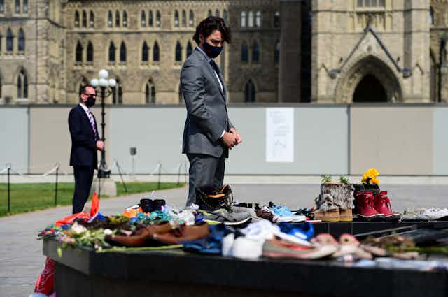Justin Trudeau, wearing a suit and a black face mask, bows his head as he looks at shoes and flowers left at the Eternal Flame on Parliament Hill