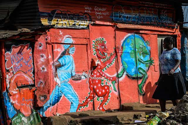 A woman walks past a mural painted on a shanty dwelling. It depicts a masked nose run-in after a devilish red virus, who is in turn running after the planet.