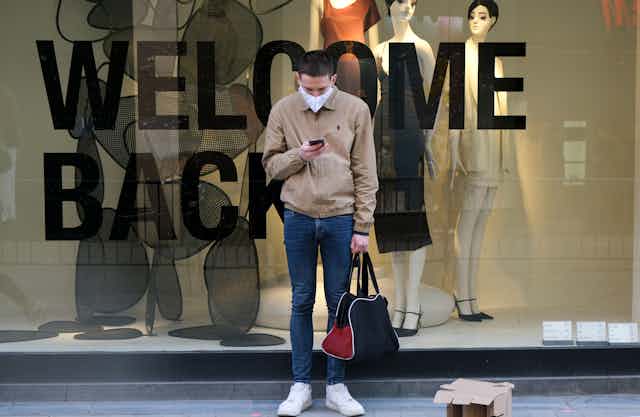 A man standing in front of a WELCOME BACK sign in a storefront window, wearing a face mask and holding a bag.