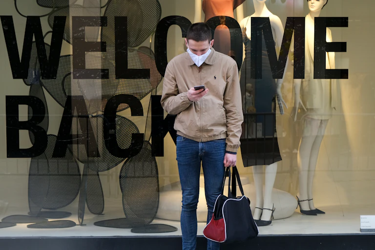 A man standing in front of a WELCOME BACK sign in a storefront window, wearing a face mask and holding a bag.