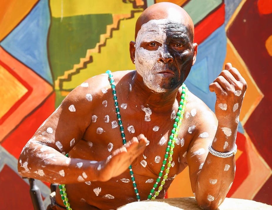 A bright traditional-patterned backdrop with a bald man in front of it beating a drum with one hand. He wears face paint - half his face white, the other black, with white paint spots covering his torso.