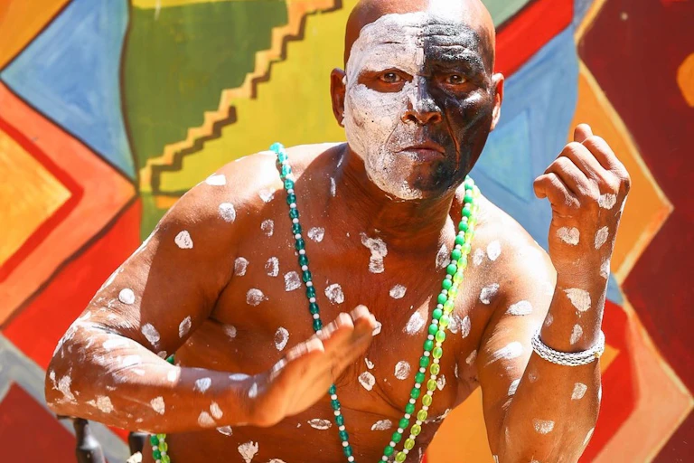 A bright traditional-patterned backdrop with a bald man in front of it beating a drum with one hand. He wears face paint - half his face white, the other black, with white paint spots covering his torso.