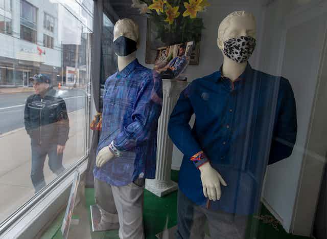Store mannequins sport face masks in a show window as a man walks by outside.