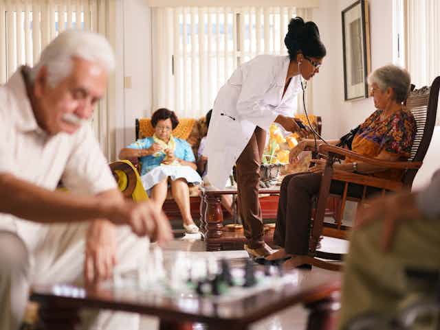 Older adults sitting in a room including a woman knitting, a man playing chess and a woman having her blood pressure checked