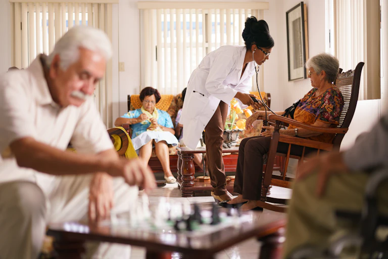 Older adults sitting in a room including a woman knitting, a man playing chess and a woman having her blood pressure checked