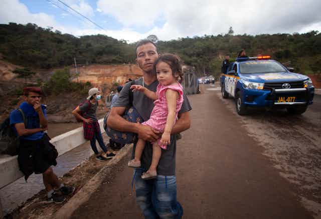 A man carries a young girl while people with backpacks sit beside a road and a police car is nearby