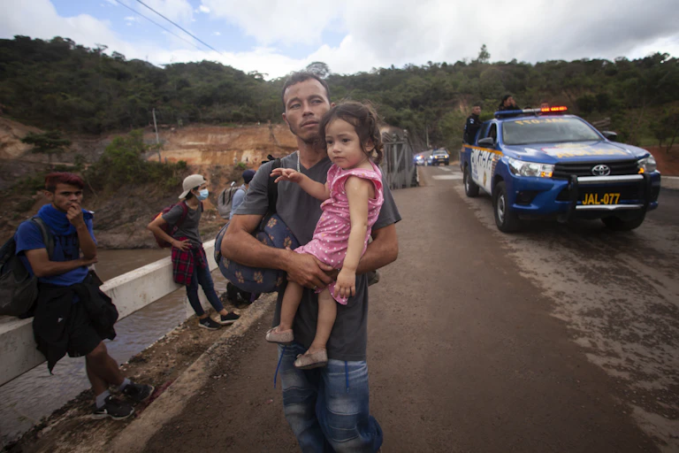 A man carries a young girl while people with backpacks sit beside a road and a police car is nearby