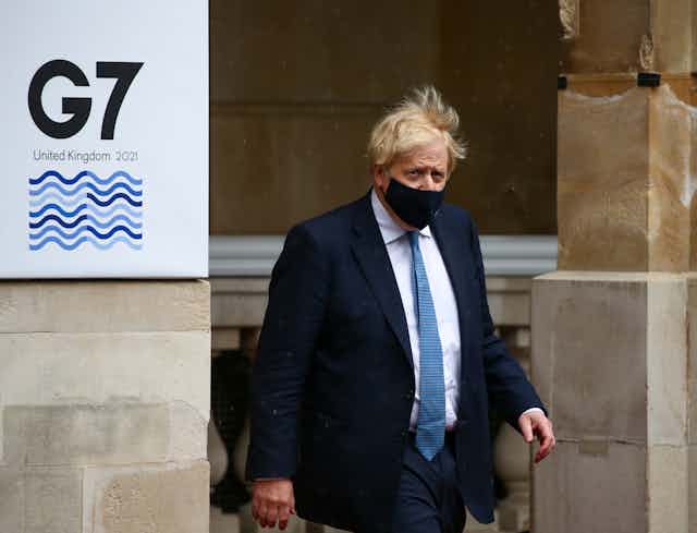 Wearing a face mask, UK Prime Minister Boris Johnson walks by a banner reading G7 UNITED KINGDOM 2021.