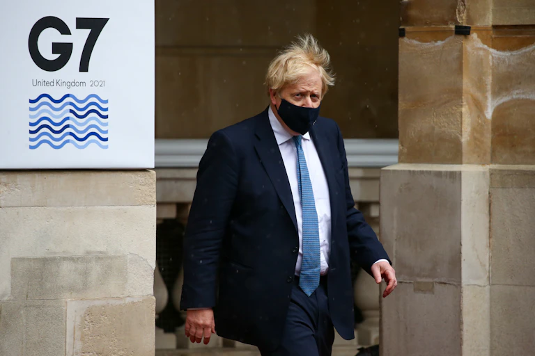 Wearing a face mask, UK Prime Minister Boris Johnson walks by a banner reading G7 UNITED KINGDOM 2021.