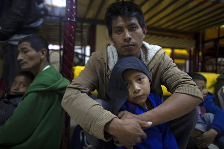 Miguel Martin sits with his 7-year-old son at a migrant shelter in Mexico with other people sitting around them.